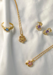 En delikat smykkekolleksjon bestående av ring, øredobber og to halskjeder med ulik farge på stenene, på em hvit bakgrunn. 