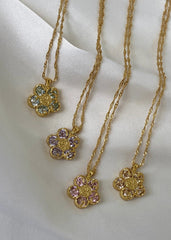 Et utvalg av gullfargede smykker med blomstermotiv og blå, lilla og klare stener, elegant presentert på hvitt underlag.