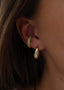 Nærbilde av øret til en kvinne med brunt hår. Hun har på seg Pace Earcuff med glitrende oliven stener, i øret henger også Dome Hoops i gull.