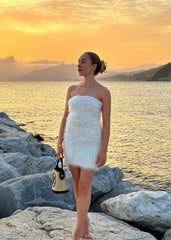 Kvinne kledd i hvit fuskepelskjole og beige kåpe, med et delikat gullkjede og anheng, poserer ved solnedgang ved sjøkanten
