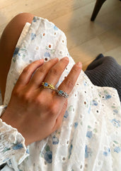 En kvinne sitter i en grålig sofa, i ført en hvit sommer kjole og poserer med sin hånd utsmykket med tre ringer og et armbånd.