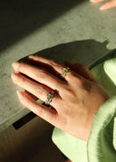 En hånd med to Melodie ringer, en ring i gull og en i sølv. Begge har oliven farget zirconia sten. Hånden lener seg på en grå marmor benk. Det er et vakkert lys fra sola som skinner på hånden. Ringene glitrer i sollyset.