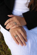 To hender som krysser hverandre over en hvit sommerkjole. På hendene har kvinnen på seg to sølvringer, melodie med oliven krystall og melodie med emerald krystall. Vakker kombinasjon av naturtoner.