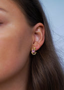 Nærbilde av en kvinne med brunt hår på en blå bakgrunn utsmykket med gulløredobb som bærer en rosa sten.