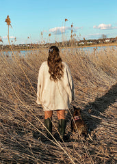 En modell står i høyt gress iført en hvit strikket genser og sorte boots. Hun har en marmorhårklype i håret