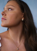 Sideprofil av en kvinne med brunt hår og hvit topp på en blå bakgrunn, utsmykket med gulløredobb og halskjede med oliven fargede stener.