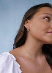 Sideprofil av en kvinne med brunt hår og hvit topp på en blå bakgrunn, utsmykket med gulløredobb og halskjede med oliven fargede stener.