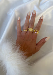 En modell med varm hudtone og beige neglelakk viser frem tre Melodie ringer og en Lumina ring i ulike farger, mot en bakgrunn av et hvitt silkestykke