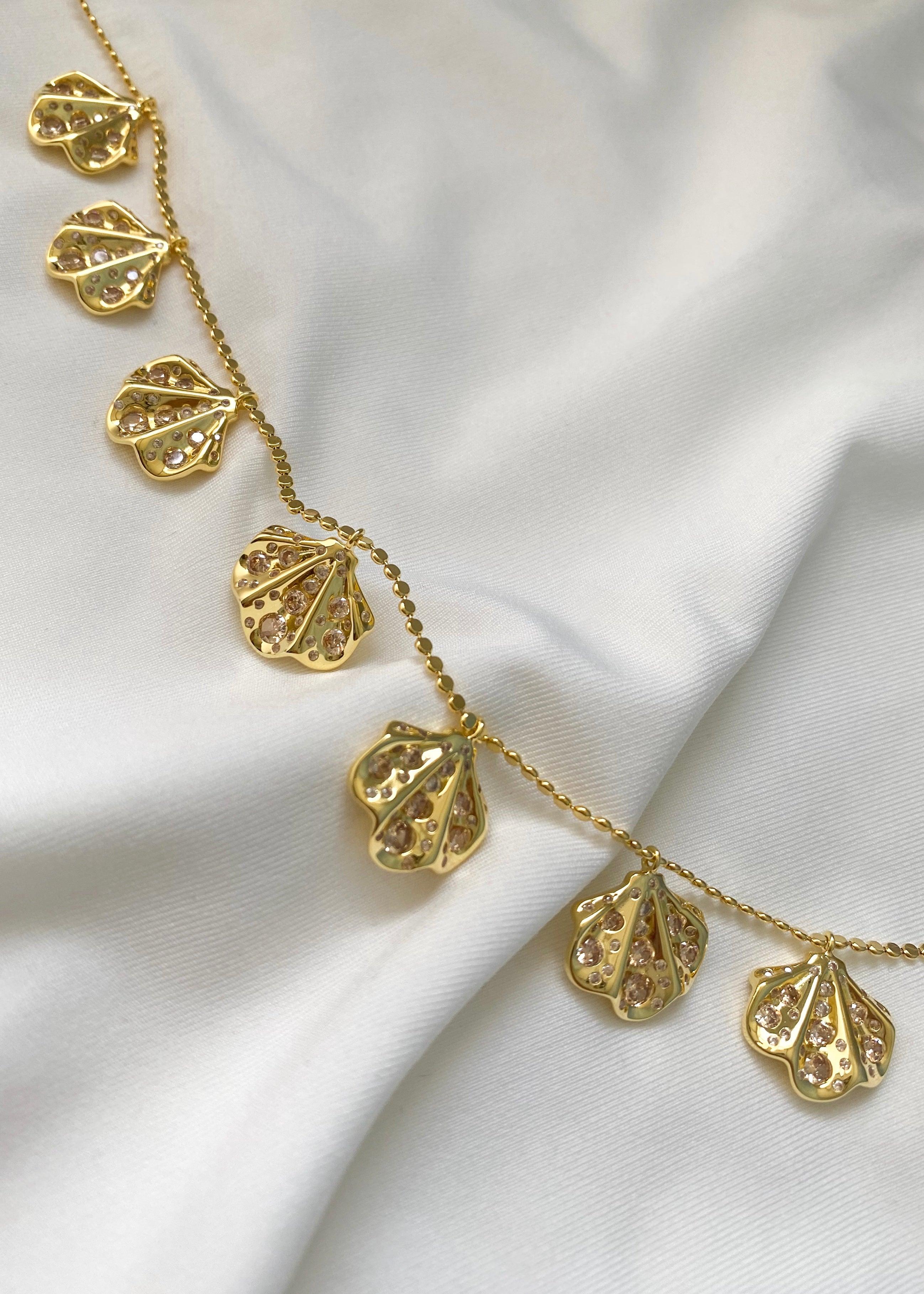 Elegant gullsmykke med kamskjell anheng som bærer champagne fargede stener på en hvit bakgrunn.