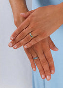 Hender med naturlige negler iført to ringer med blå firkantede steiner, én på hver hånd, mot en lys blå bakgrunn. Elegant og fargerik smykkestil.