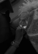 Nærbilde av en kvinnehånd som holder et glass. Kvinnen sitter og lener hånden sin inntil låret sitt. Hun har på en kjole og glitrende strømper. På armen har hun en delikat klokke og connection armbåndet plassert like ved.