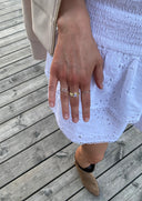 En kvinnehånd lener seg inntil en hvit sommerkjole. På hånden har hun en Melodie ring i gull med lavendel farget zirconia sten. På de andre fingrene har hun små, tynne, delikate sølvringer. Melodie ringen skiller seg ut og fanger mye oppmerksomhet med sin unike tekstur og glitrende utseende.