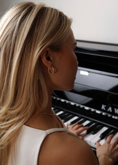 En kvinne med lyst blondt hår sitter foran en piano. Håret hennes ligger delikat bak øret, i øret har hun en vakker Dome Hoop i gull. Rundt halsen har hun et tynt gullhalskjede.