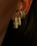 Nærbilde av en kvinne sitt øre i solnedgangen. Hun har på seg to Pave Earcuff, en med hvite glitrende zirconia stener og med oliven grønne glitrende stener. Kvinnen har også Diamond rectangle øredobber, en i Large og en i small.