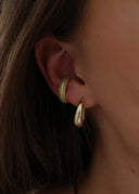 Nærbilde av øret til en kvinne med brunt hår. Hun har på seg Pace Earcuff med glitrende oliven stener, i øret henger også Dome Hoops i gull.
