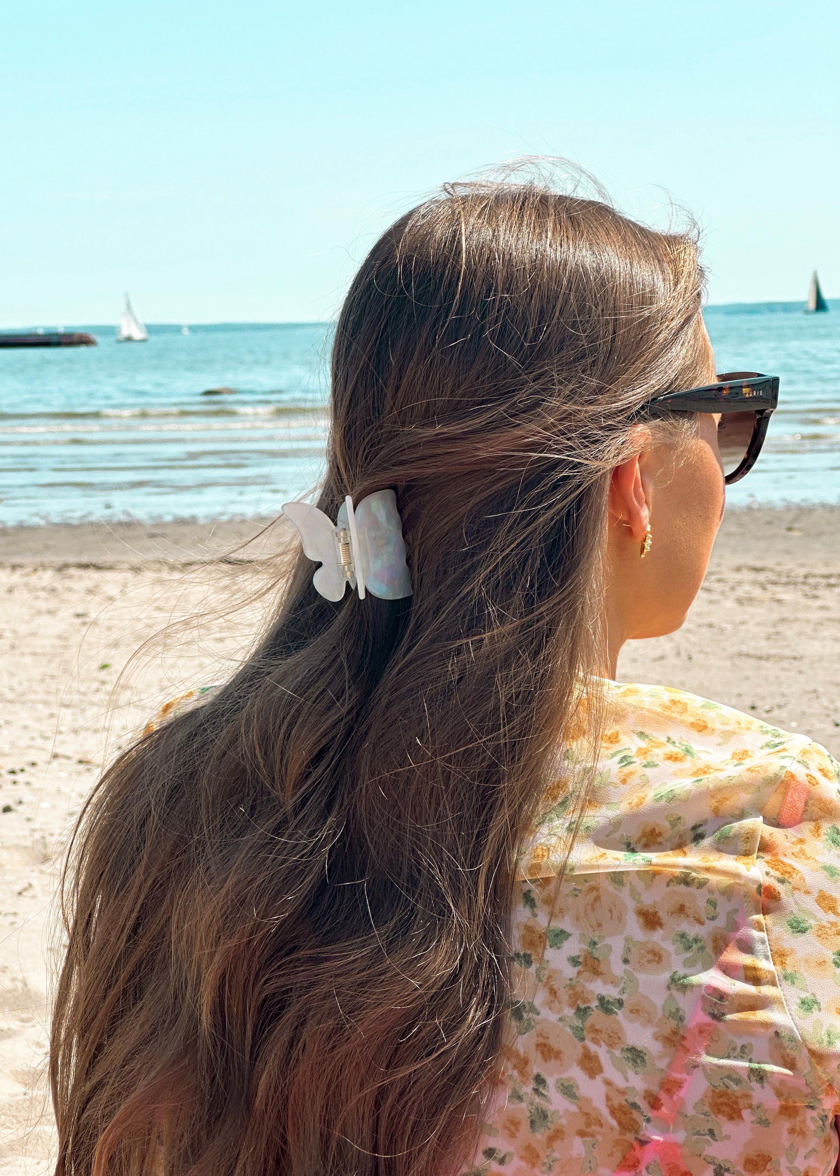 En kvinne på stranden nyter solen iført en solfylt, gul sommerkjole. Hun har langt brunt hår og bruker solbriller for å beskytte øynene sine. Håret er pyntet med en hvit sommerfuglhårklype, som gir et delikat og sommerlig preg.