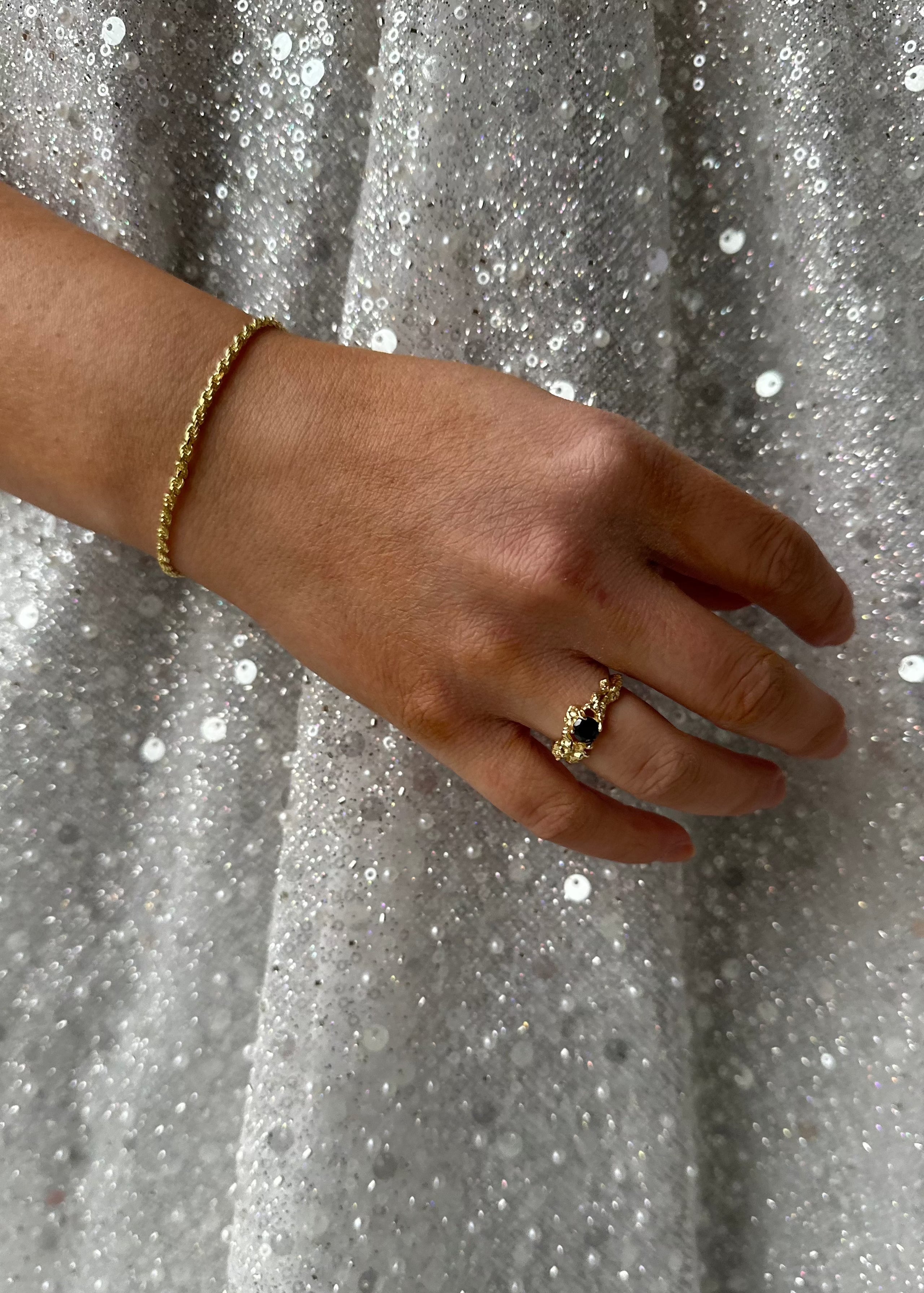 Kvinnehånd med gullarmbånd og elegant sort steinring mot en glitrende sølvbakgrunn for luksuriøse smykker.