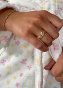 En hånd som lener seg inntil en blomstrende badekåpe med en melodie ring i gull med lys rosa sten.  På armen har hun et stjernestøv armbånd i gull.