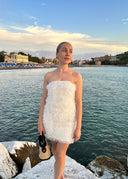 Stilig kvinne i hvit off-the-shoulder kjole poserer med smykker ved solnedgang, med utsikt over sjøen