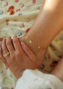 Nærbilde av en kvinne ankel som har på seg Sunflower Anklet i gull. Kvinnen holder en hånd på foten sin, på hånden har hun på seg en matchende Star Wave ring. De små delikate solsikkene gir en sommerlig og frisk look.