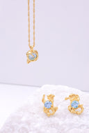 Lykkesten halskjede med blå cubic zirconia sten  og matchende melodie øredobber med blå stein er plassert på en delikat hvit bakgrunn. Gullfargen på smykkene skinner på bildet og skaper et elegant intrykk.