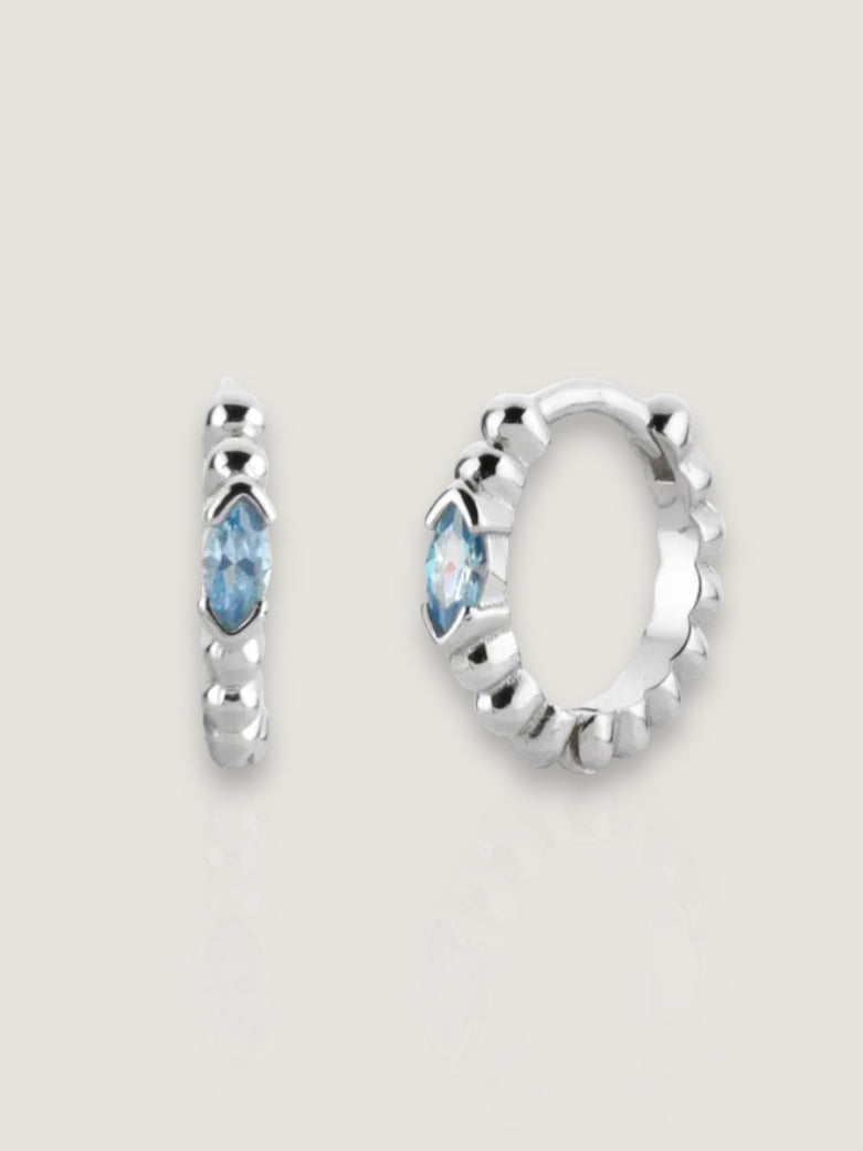 På en beige bakgrunn ligger twisted eye hoops i sølv med en blå sten. Disse sølvoresmykkene har en vridde design og gir en elegant touch med den blå stenen som en fokuspunkt.