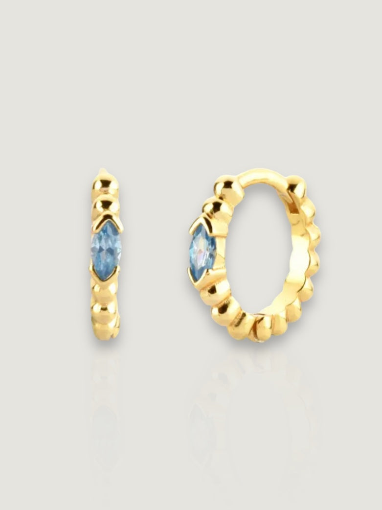 På en beige bakgrunn ligger twisted eye hoops i gull med en blå sten. Disse øreringene har en vridde design og gir en elegant touch med den blå stenen som en fokuspunkt..