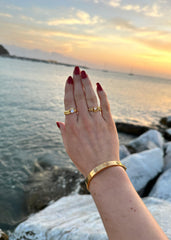 En kvinne poserer handen sin utover en vakker sjøutsikt i ført rød neglelakk og diverse gullsmykker.